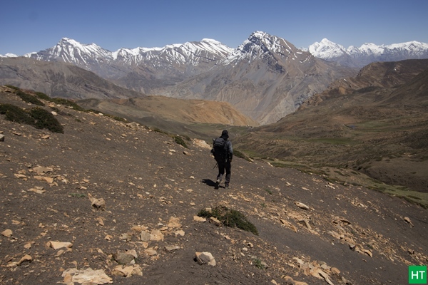Trekker on high, barren slopes above Spiti Valley with snow‑capped peaks ahead — the village‑to‑village, on‑foot way to see Spiti