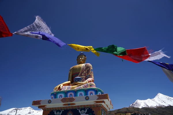 Langza’s Buddha statue under prayer flags, snow peaks behind — a high Spiti village (~4,200 m) many road trippers visit.
