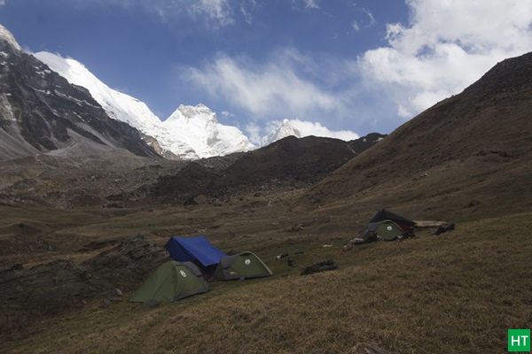 Wide open campsite at Kedar Kharak with tents pitched on grass, snow peaks visible in background